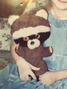 Old family photo of a child in a blue dress hugging a brown childhood raccoon plush