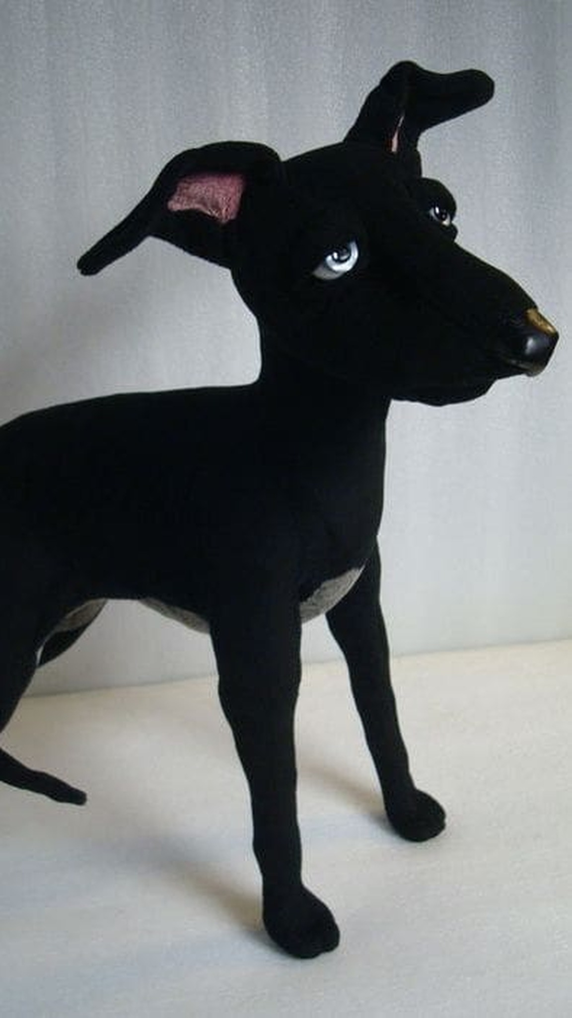 Black whippet stuffed animal standing, blue glass eyes and a slim body, a pet replica plush made to order