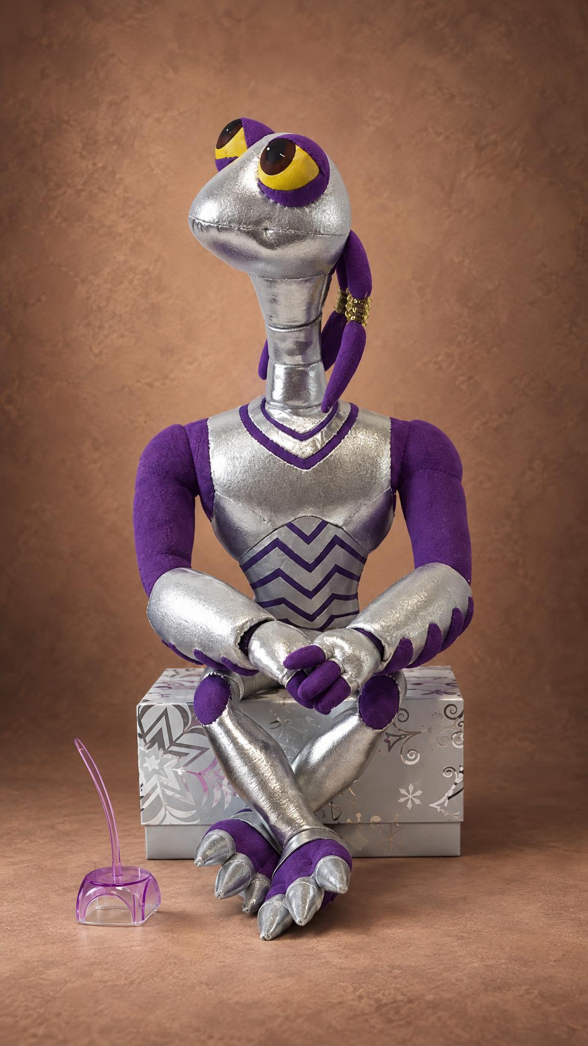 Handmade Croakagy alien plush sitting cross-legged on a silver gift box, yellow eyes and purple tendrils