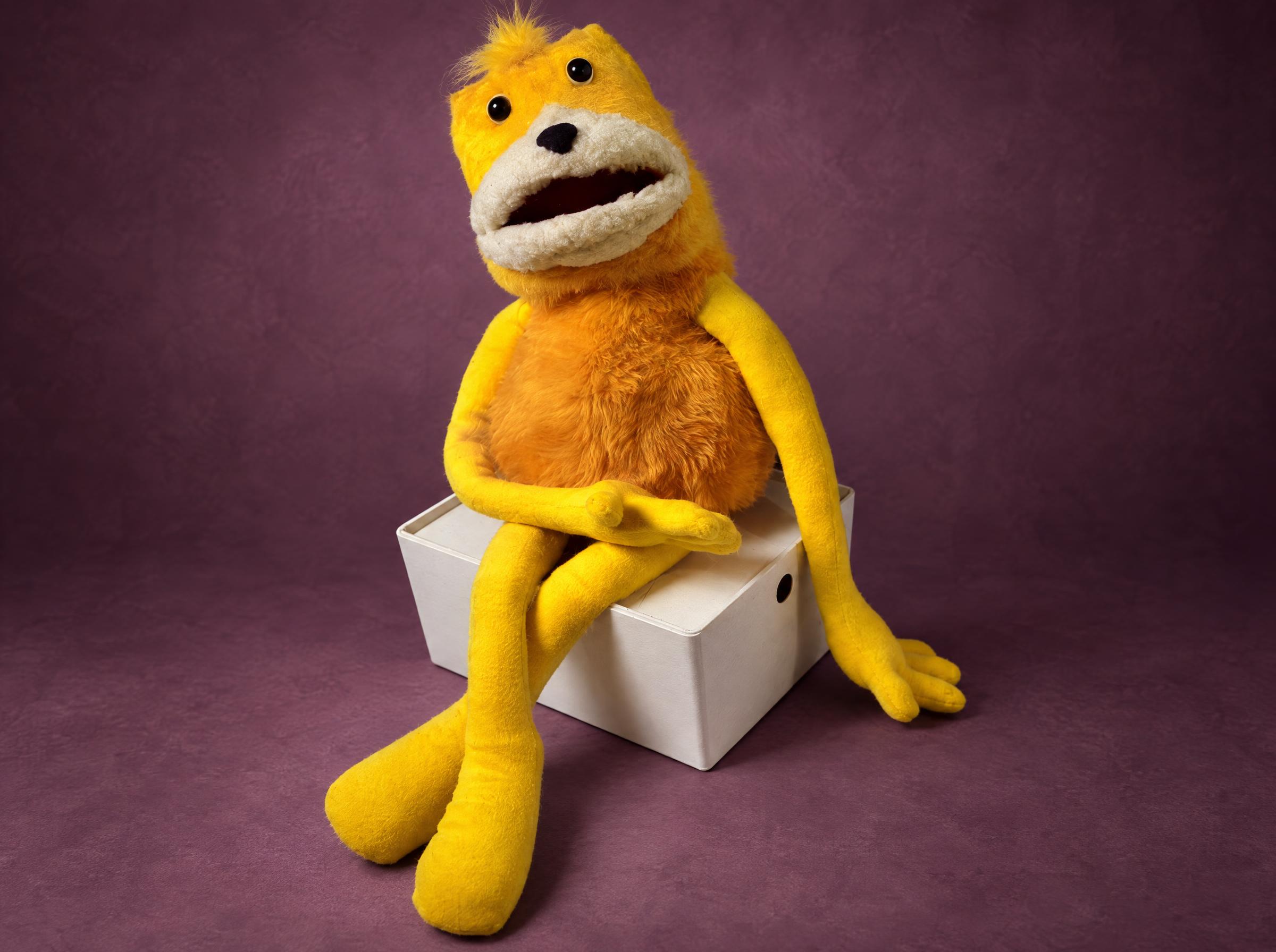 Handmade Flat Eric puppet made to order, yellow fur, long floppy arms, sitting on a white box