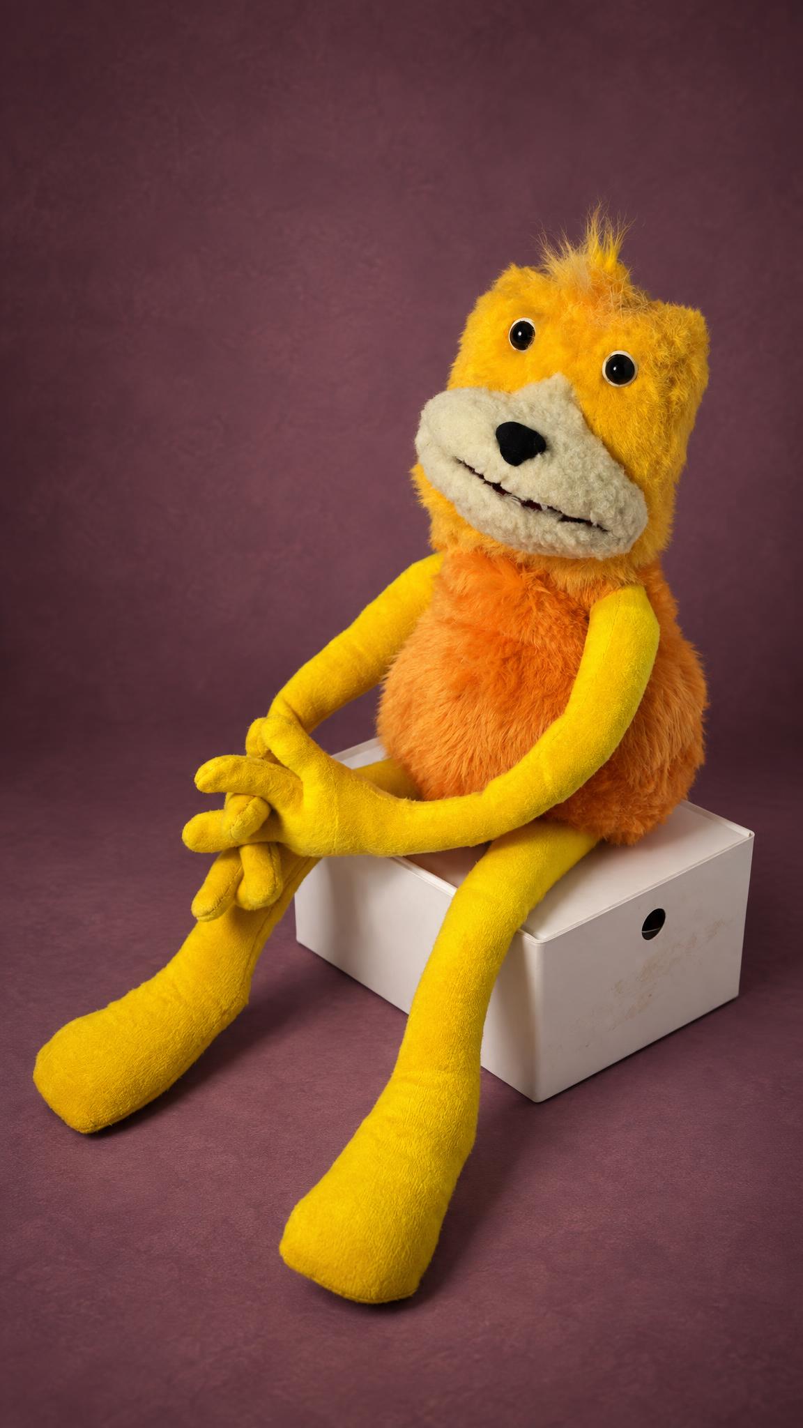 Flat Eric puppet handmade to order, yellow hands folded on his knee, orange fur body, sitting on a white box