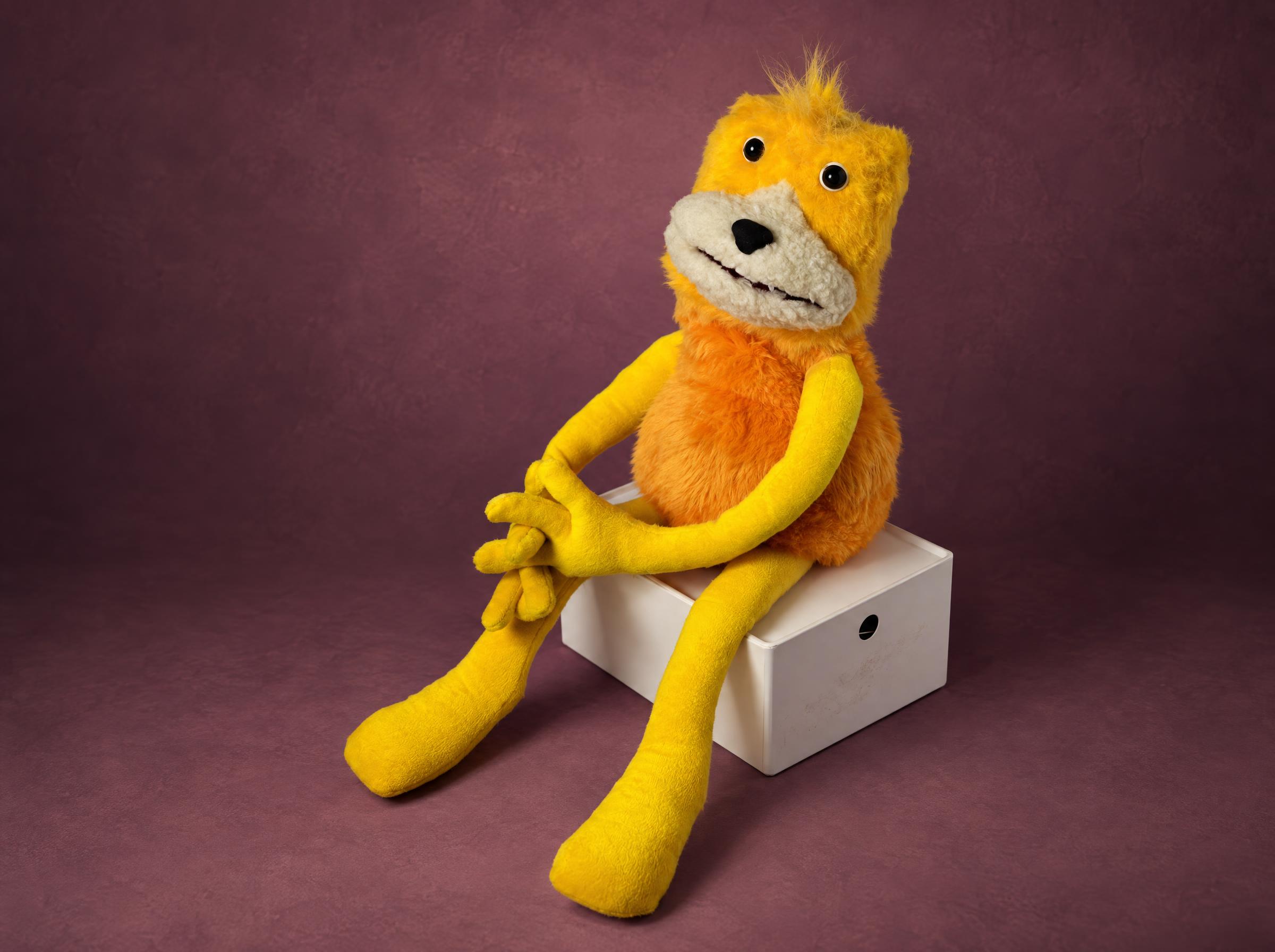 Flat Eric Puppet with Folded Hands, Handmade Flat Eric puppet handmade to order, yellow hands folded on his knee, orange fur body, sitting on a white box