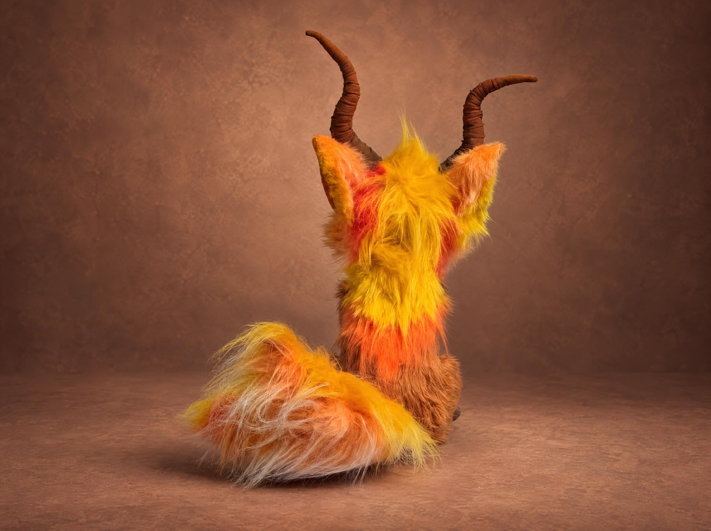 Horned fox plush from behind, mane and horns Back of the horned fox plush: yellow and orange faux-fur mane, curved brown horns and a fluffy tail, custom made to order