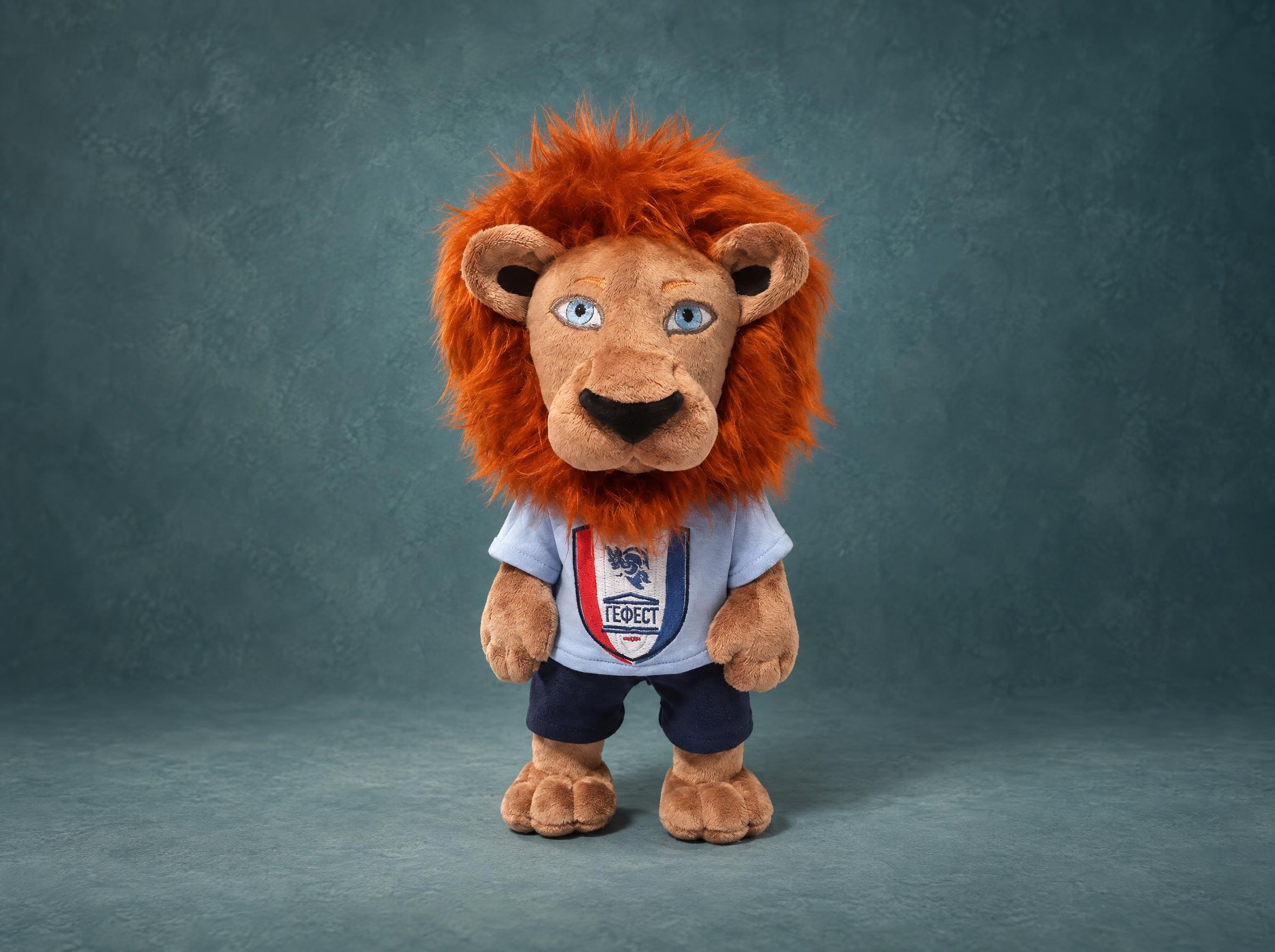 Hugo, a lion mascot plush for FC Gefest: orange faux-fur mane, crest T-shirt, navy shorts, handmade to order