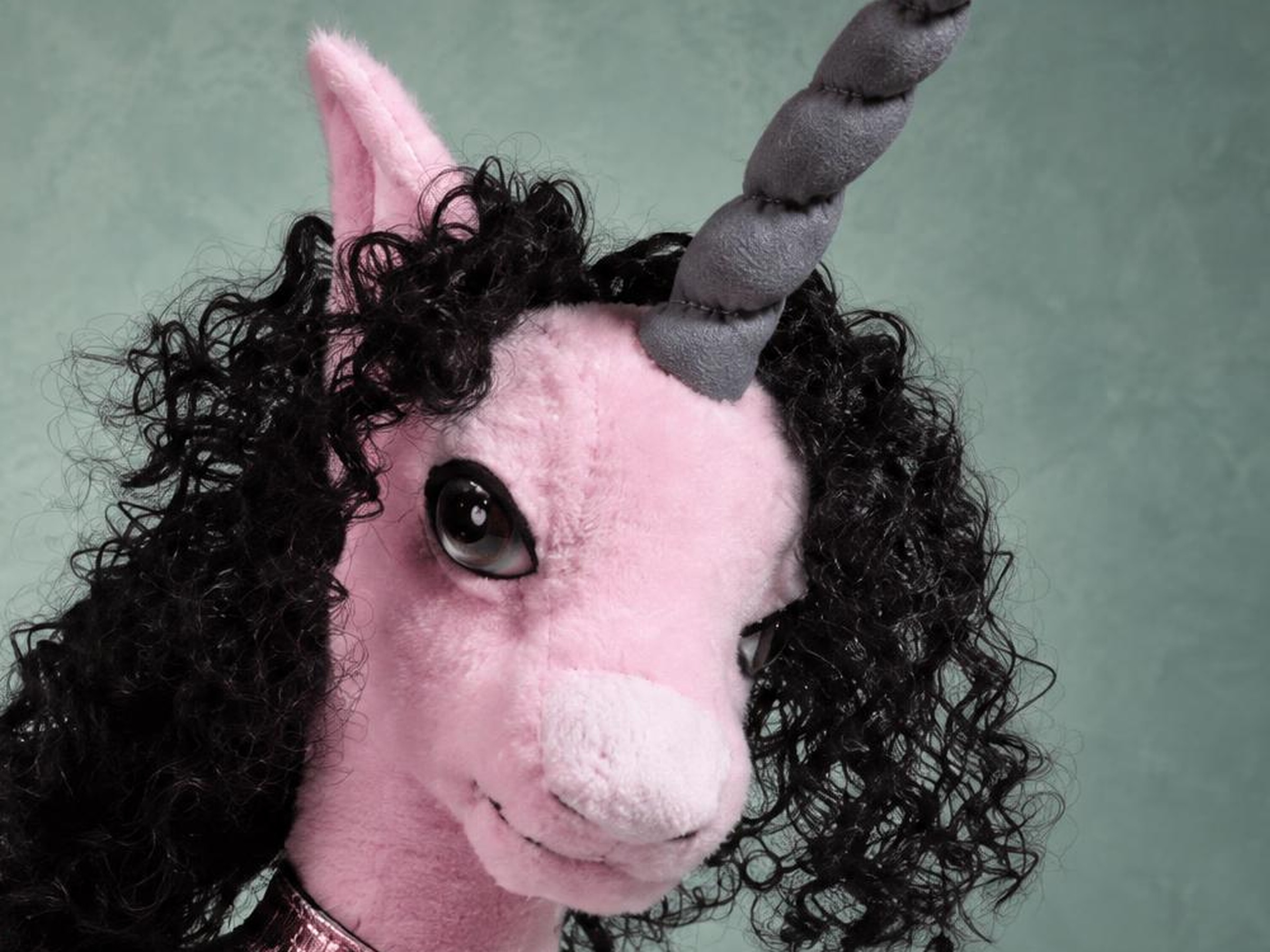 Handmade pink unicorn plush with a black curly mane Handmade pink unicorn plush face with a glossy dark eye, a grey spiral horn and a mane of black curls