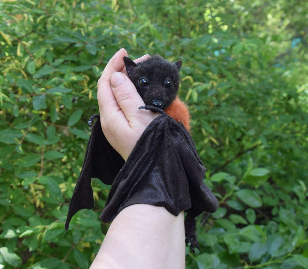 Palm-sized bat plush Palm-sized bat plush curled in a hand, wings wrapped around the wrist, a realistic fruit bat sewn to order