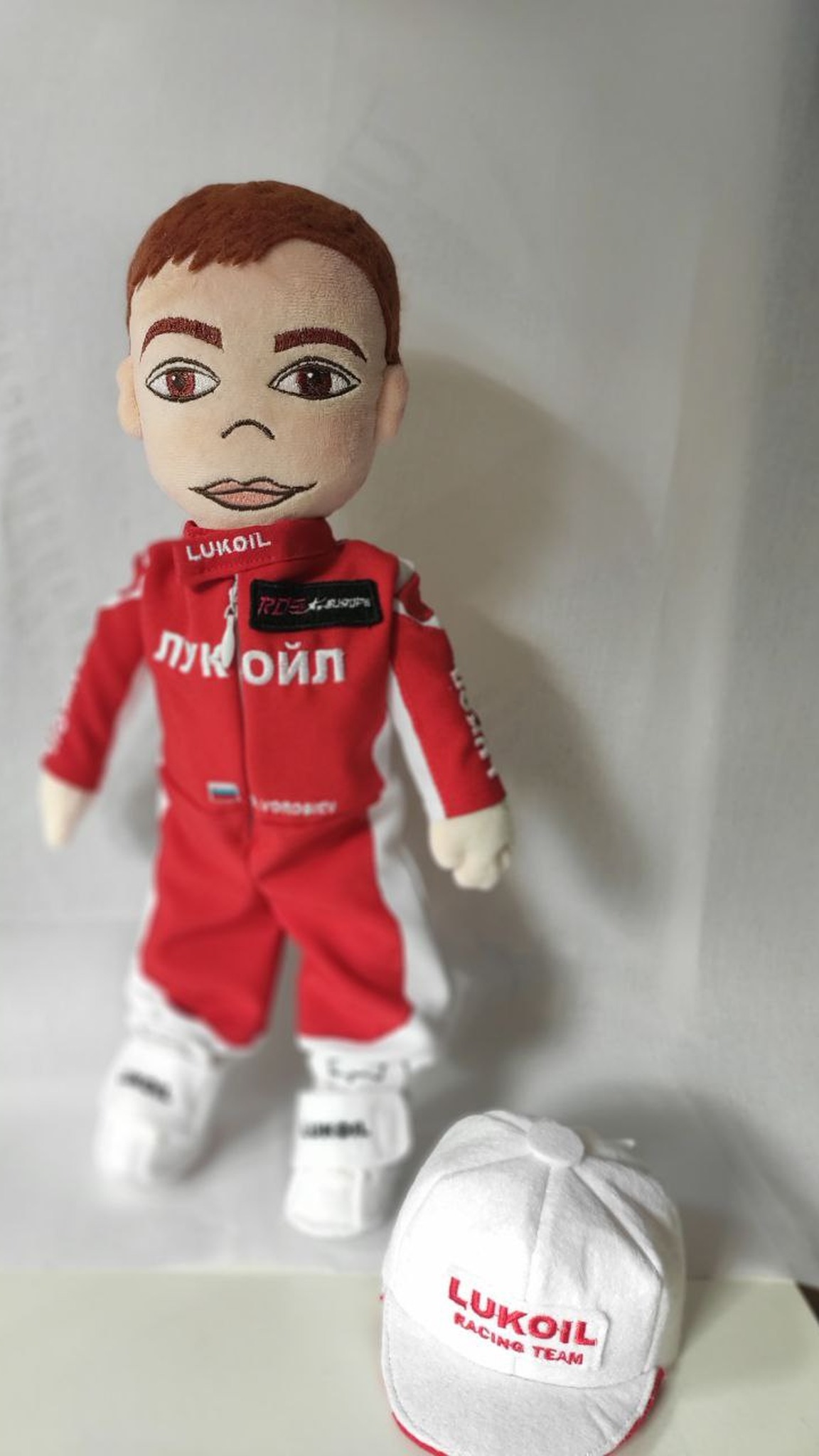 Branded plush doll with short hair and a removable team cap beside him, custom corporate gift made to order