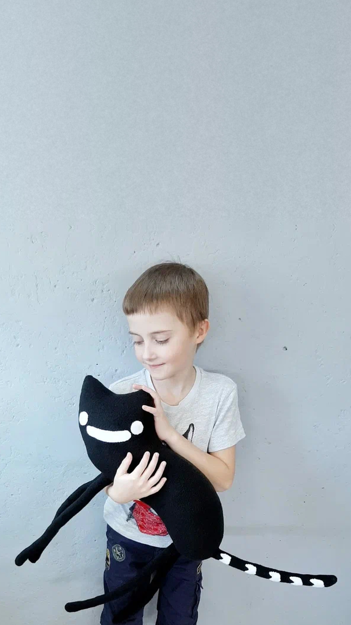Boy hugging a custom black cat plush with a white line eye and smile, made to order from an illustration