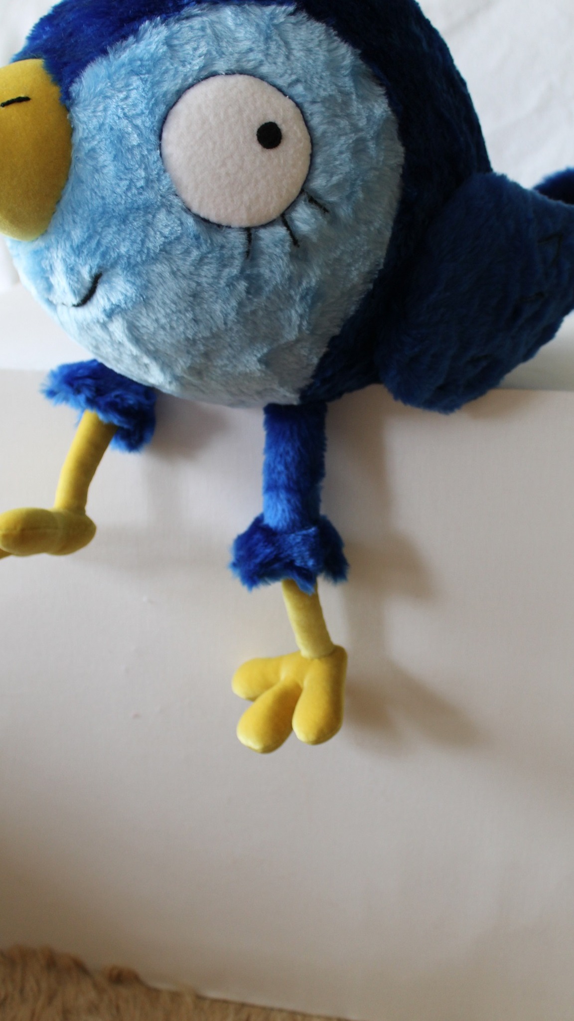 Custom blue bird plush sitting on a ledge with its yellow legs hanging down, embroidered lashes and smile