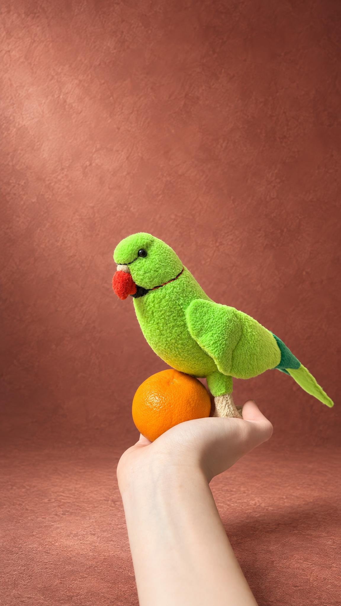 Green ringneck parrot plush sewn to order as a copy of a pet bird, red beak and long tail, standing on a hand