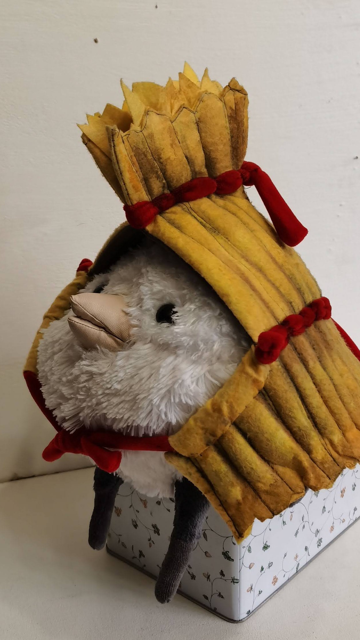 Fluffy bird plush under a ribbed straw cape with a fringed tuft and red velvety bows, sewn by hand to order
