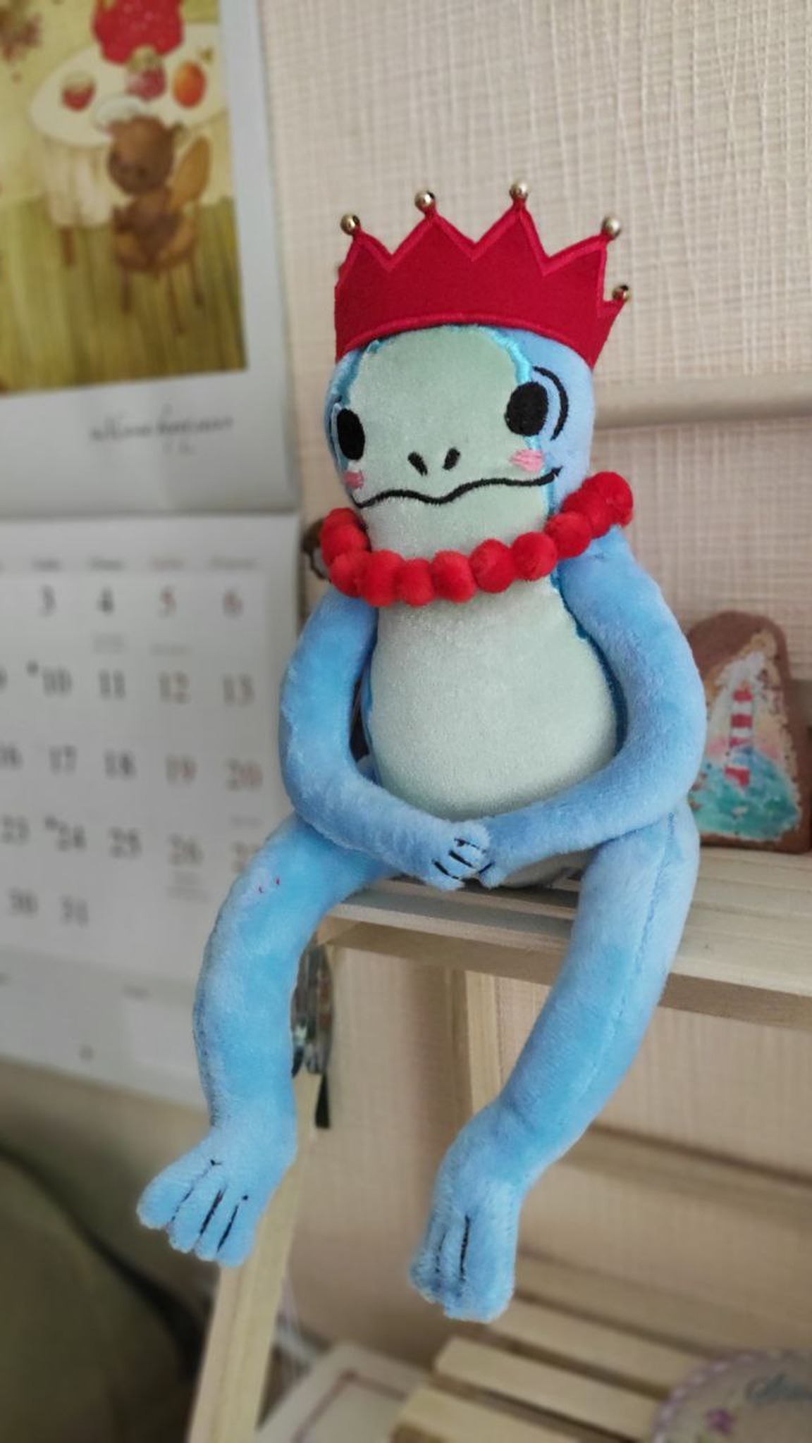 Frog princess plush sitting on a shelf edge, hands folded, red crown and pom-pom necklace, made to order