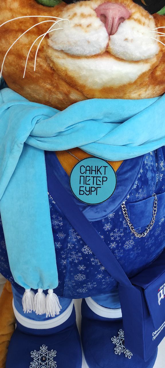 Giant cat plush scarf, badge and pocket chain Giant cat plush details: light-blue scarf with white yarn tassels, round badge and a chain on the pocket
