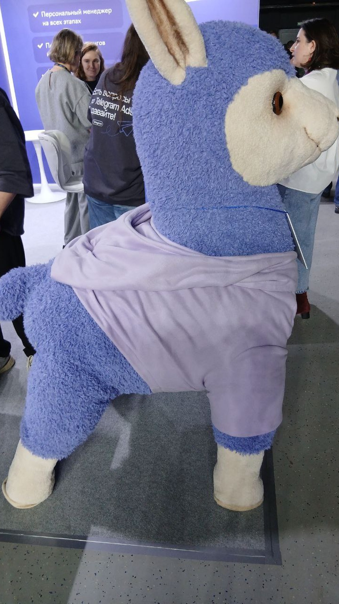 Giant llama plush mascot from behind: blue curly plush legs and tail, lilac hoodie, cream hooves, handmade