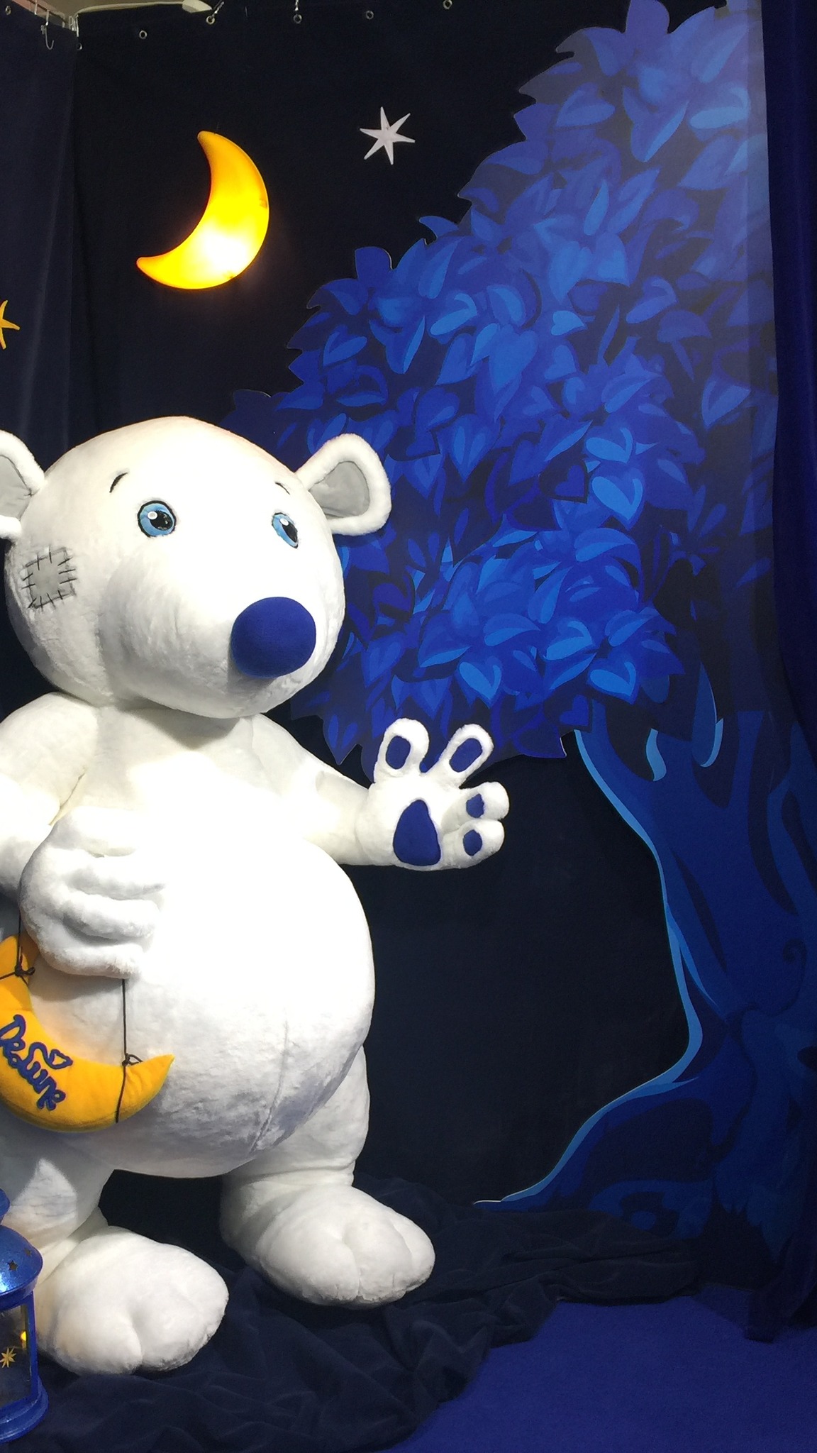 Giant teddy bear mascot by a painted blue tree at an exhibition booth, custom plush made from brand art