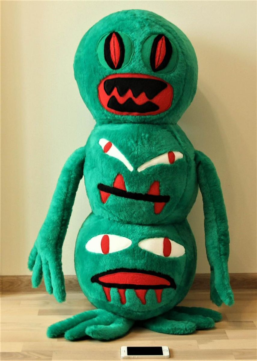 Big green monster plush with a phone for scale Big handmade green monster plush from a drawing standing by a wall, a phone at its feet for scale