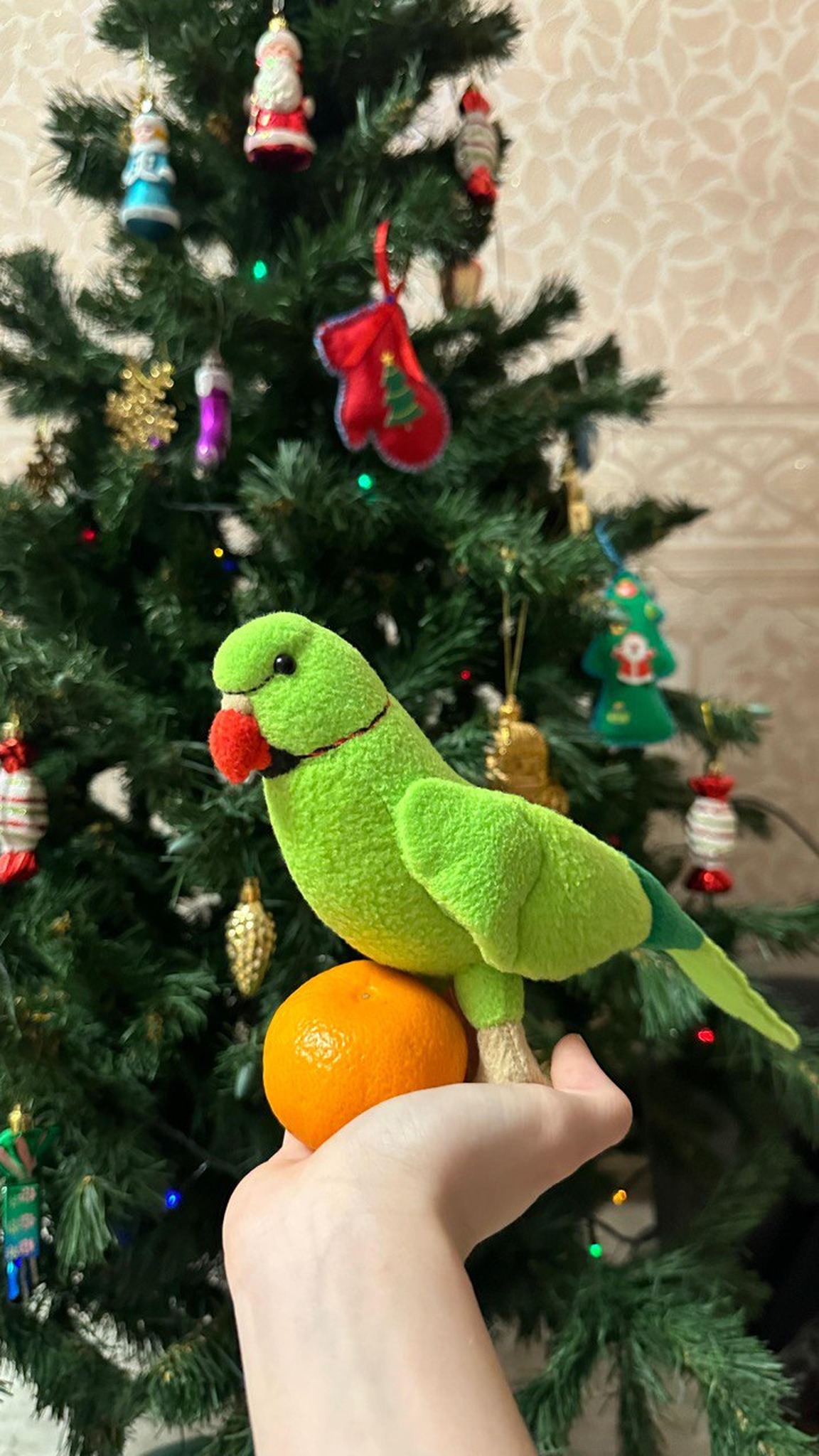 Custom green parrot plush with red beak and neck ring on a hand by a New Year tree, sewn to order