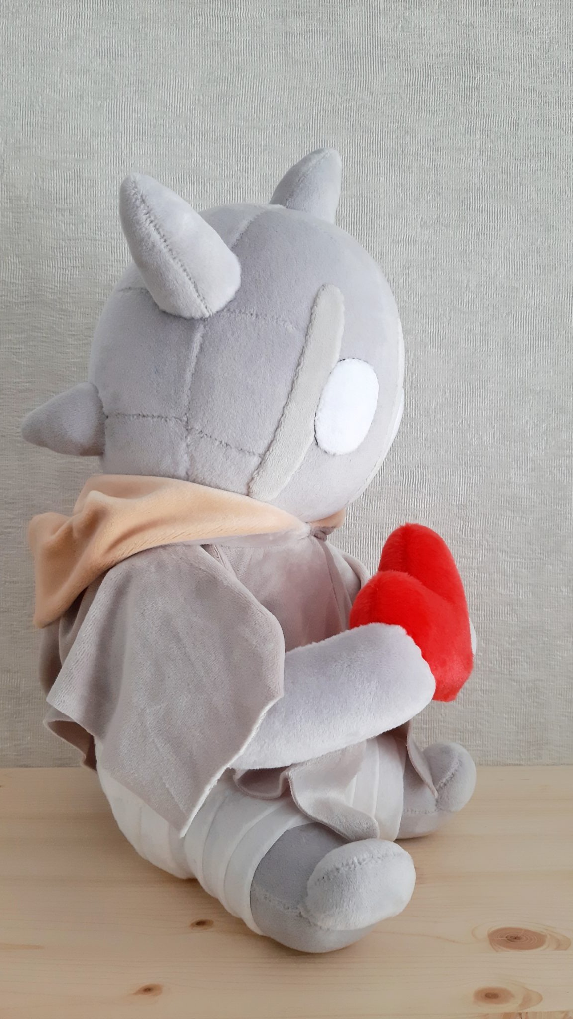 Horned monster plush in a grey cloak clutching a red heart, mask-like stitched face and white eye, custom made