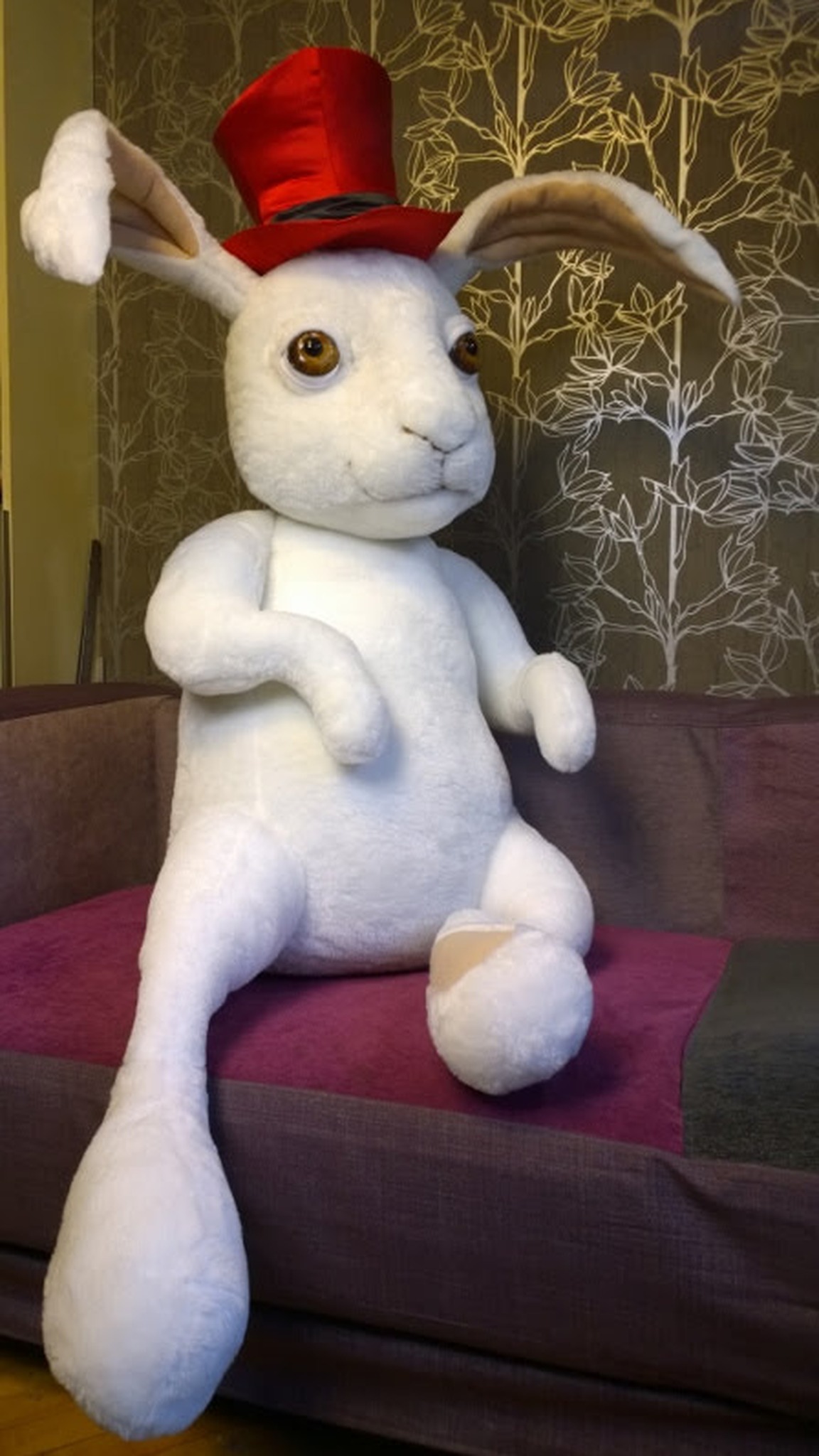 Large rabbit stuffed animal sewn to order, sitting on a sofa with legs hanging over, red top hat, amber eyes