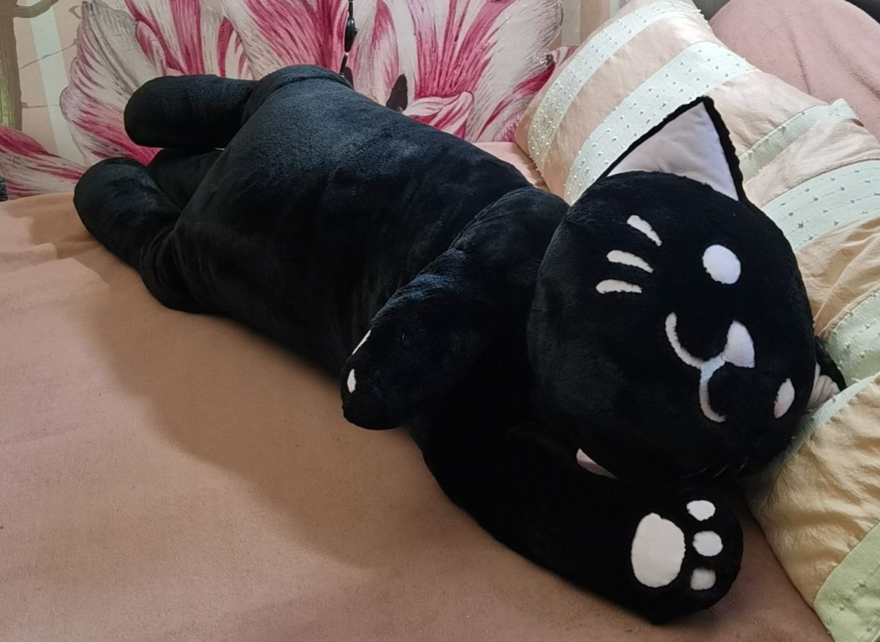 Long black cat plush cuddle pillow Long black cat plush cuddle pillow made to order, lying on a bed pillow with one paw raised