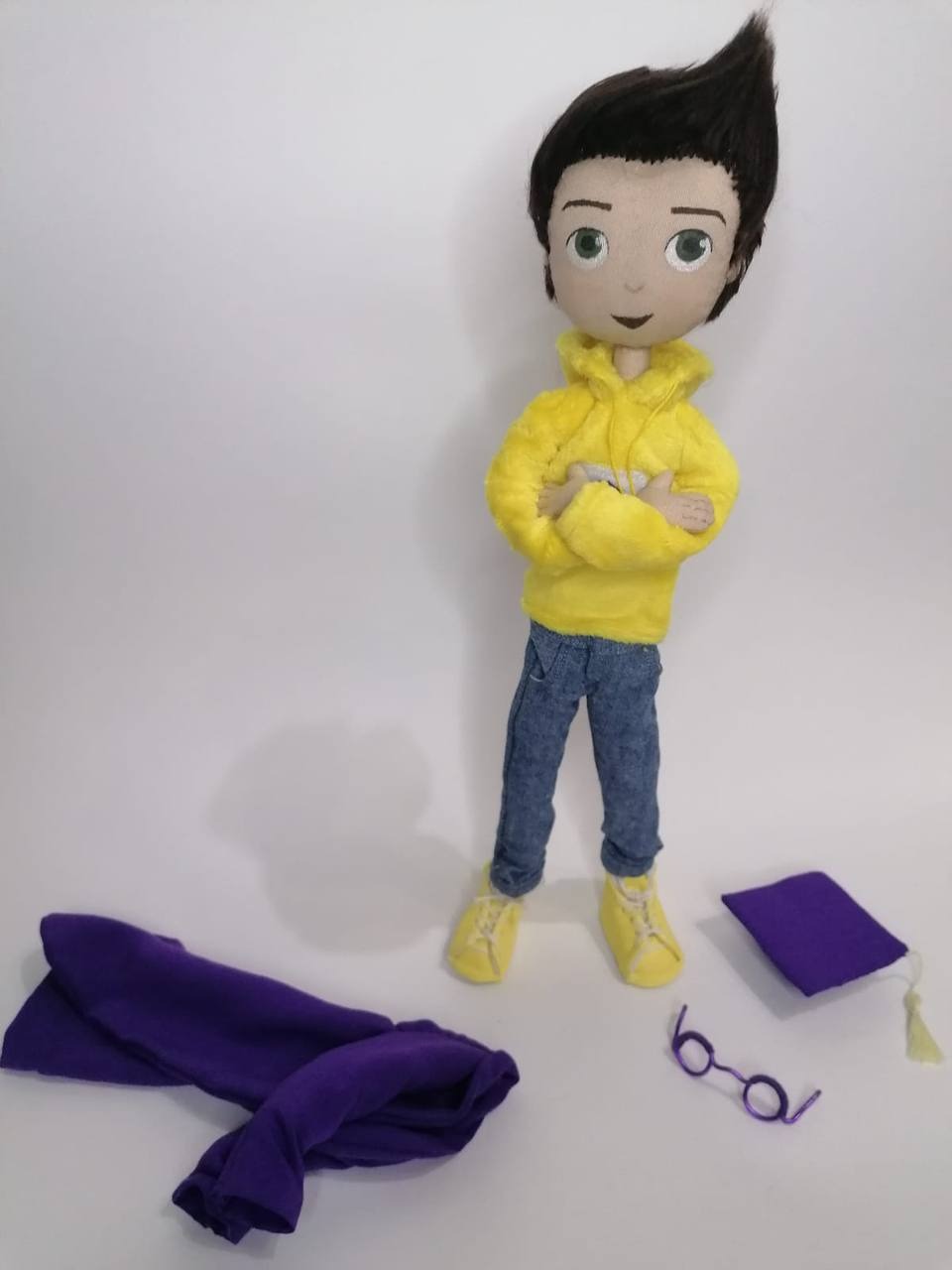 Mascot doll with removable gown, cap and glasses Graduate mascot doll with arms crossed, his removable violet gown, cap and wire glasses beside him, handmade