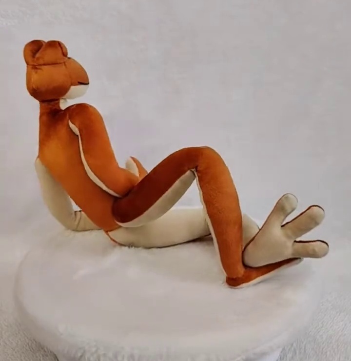 Patila frog plush lounging Patila frog plush lounging with long orange legs and a cream foot raised, handmade amphibia sapiens toy