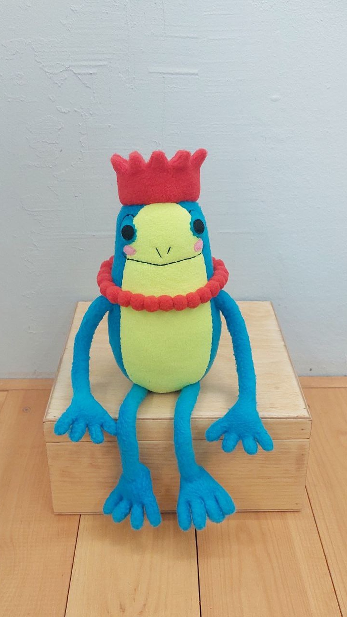 Turquoise frog plush in a red crown sitting on a wooden box, long legs down, made to order from a sticker